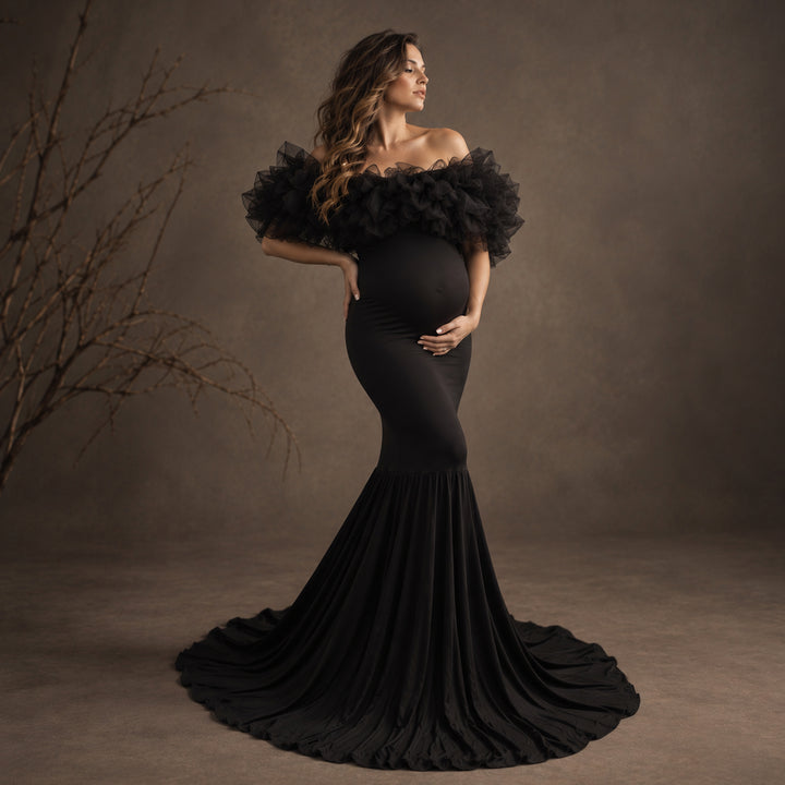 A pregnant model wearing a black off-the-shoulder maternity gown with dramatic ruffled sleeves and a mermaid-style floor-length skirt.