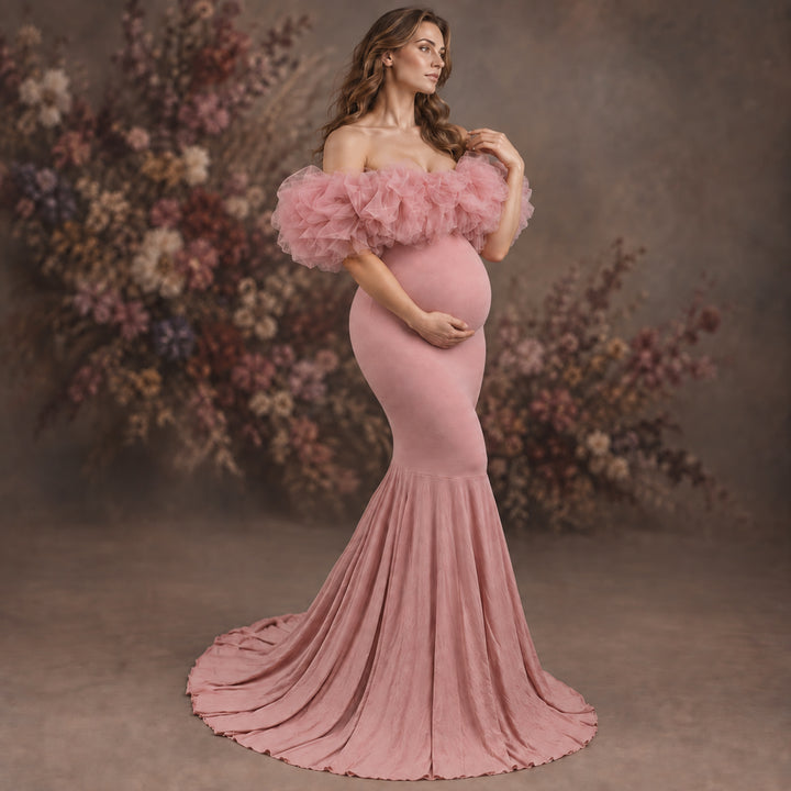 A pregnant model wearing a dusty pink off-the-shoulder mermaid gown with a ruffled tulle bodice and a long flowing skirt, posed in front of a soft-focus floral studio backdrop.