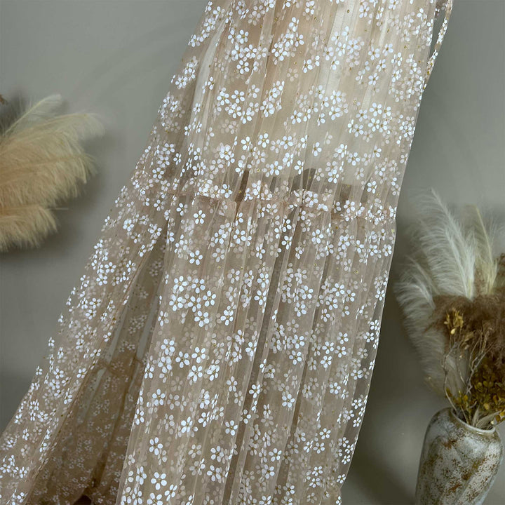 A khaki tulle dress with white floral embroidery and gold glitter accents, displayed against a neutral backdrop with dried floral arrangements.