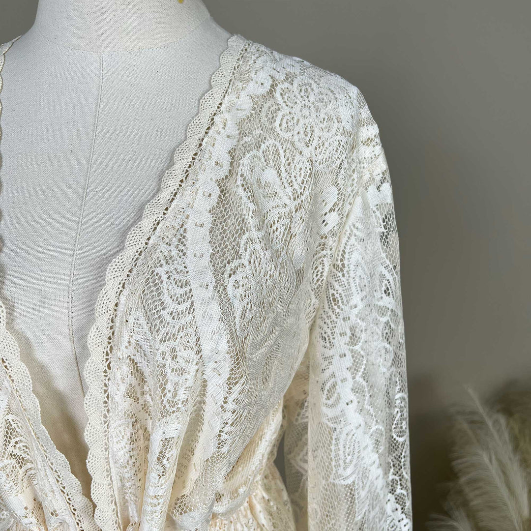 Close-up of a beige lace displayed on a mannequin. The image shows the shoulder, long sleeve, and V-neckline with scalloped lace edging.