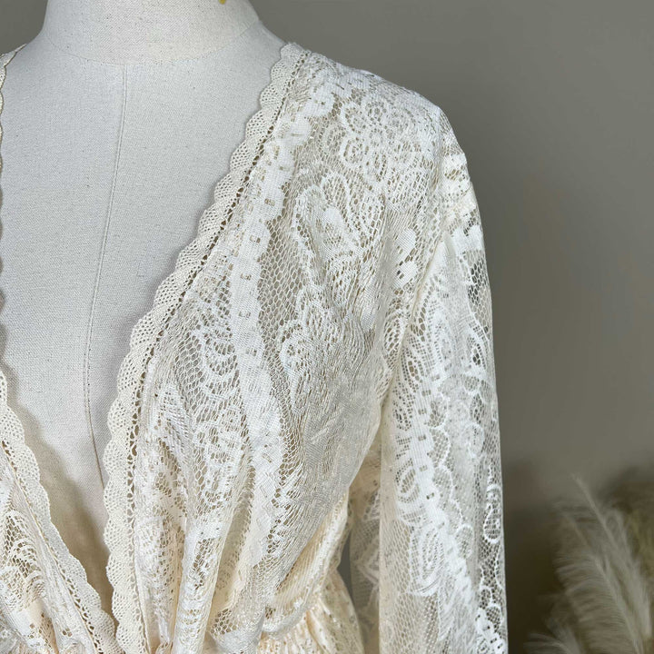 Close-up of a beige lace displayed on a mannequin. The image shows the shoulder, long sleeve, and V-neckline with scalloped lace edging.