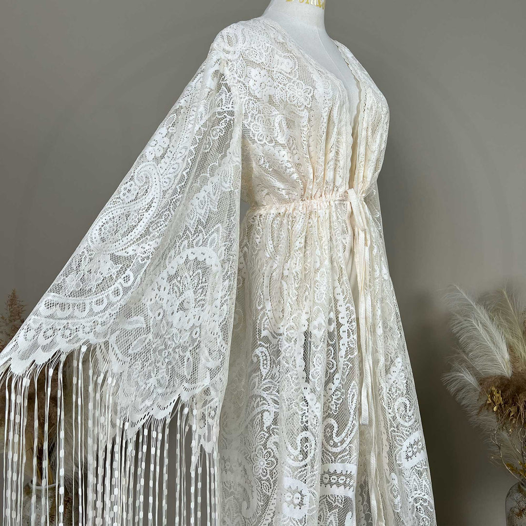 A beige lace with bell sleeves and fringe trim, displayed on a mannequin. The garment features a V-neckline, an elasticated waist with a tie closure.