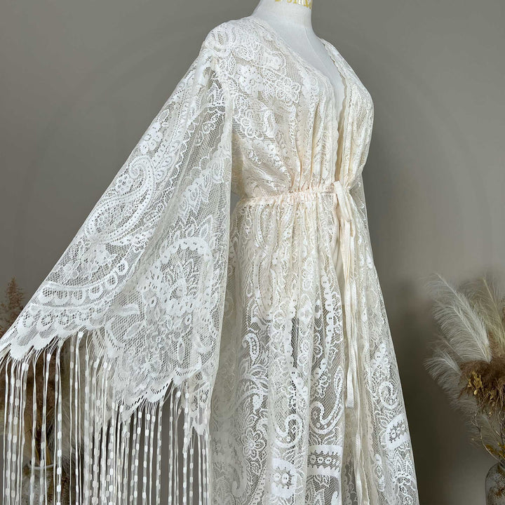 A beige lace with bell sleeves and fringe trim, displayed on a mannequin. The garment features a V-neckline, an elasticated waist with a tie closure.