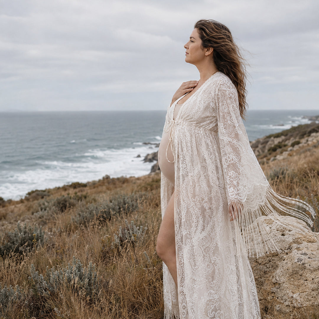 A pregnant woman stands on a grassy hillside near the ocean coast, wearing a beige lace open-front robe with bell sleeves and fringe trim.