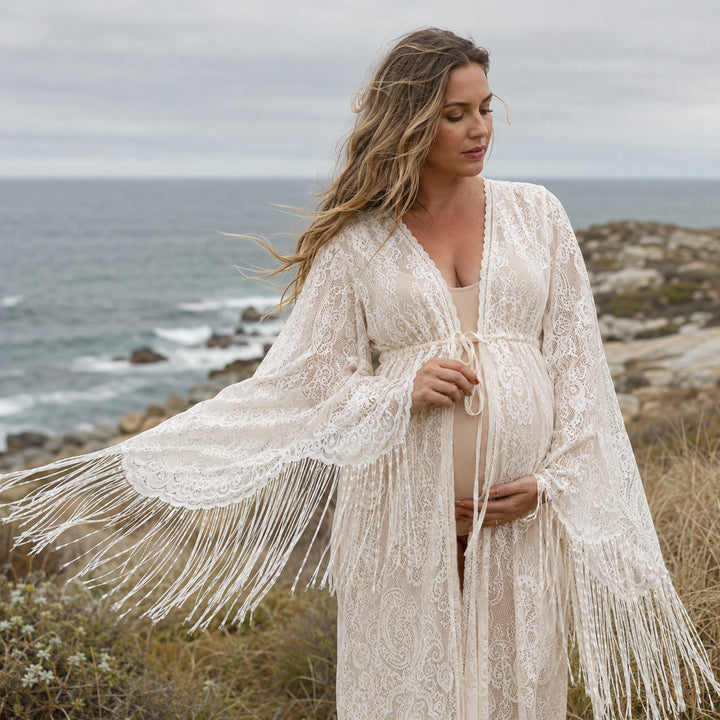 A pregnant woman stands on a grassy coastal cliff overlooking the ocean, wearing a beige lace maternity with long bell sleeves and fringe trim. 