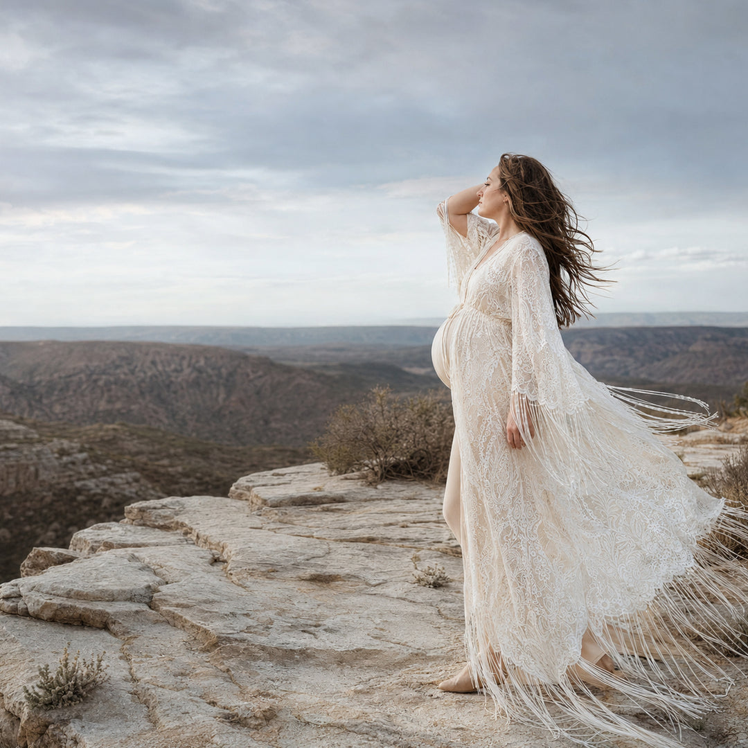 A pregnant woman stands in profile on a rocky cliff edge with a vast canyon landscape in the background, wearing a floor-length beige lace robe with fringe details.