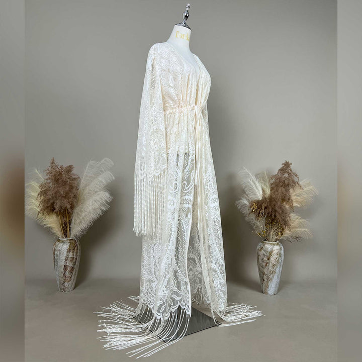 A beige lace maternity robe with long sleeves and fringe trim, displayed on a mannequin. The robe features an open front design, a V-neckline, and a floor-length hem with dangling fringe details extending outward.
