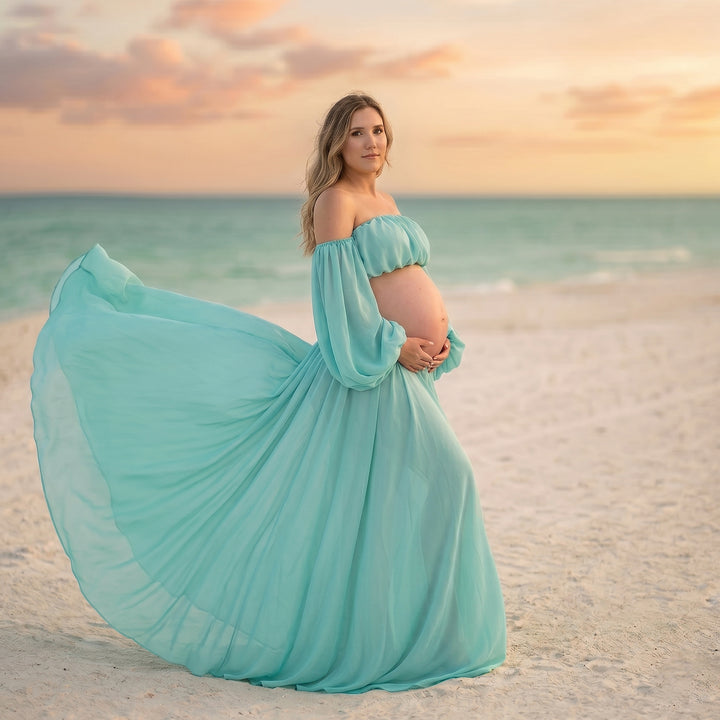 A pregnant woman stands on a beach wearing a Gaia mint flowy chiffon off-shoulder dress, holding her belly。