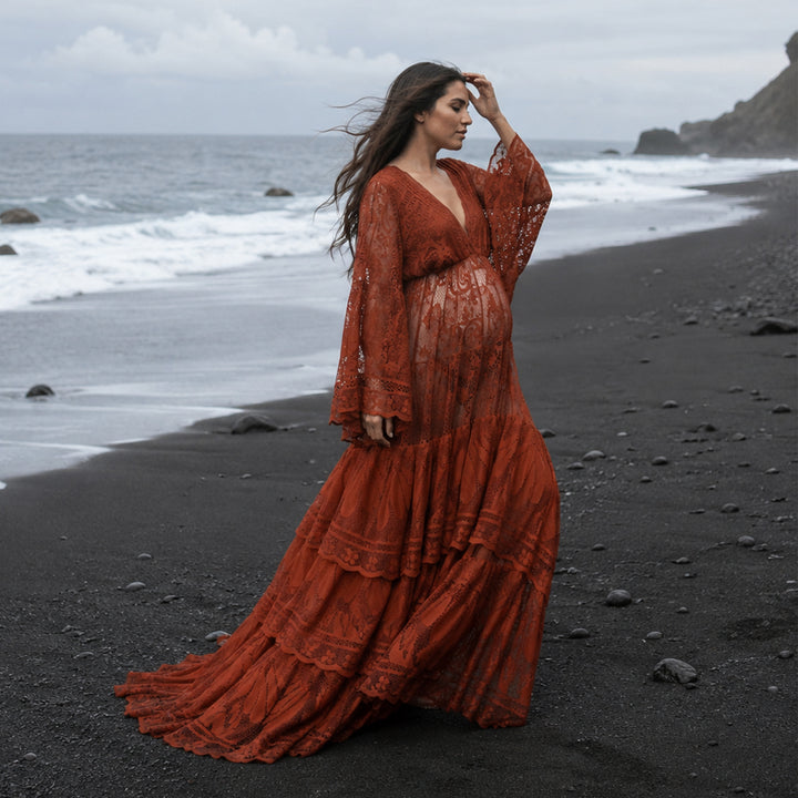 A pregnant woman stands in profile on a rocky black beach near the water's edge, wearing a long terracotta lace dress with long sleeves. 