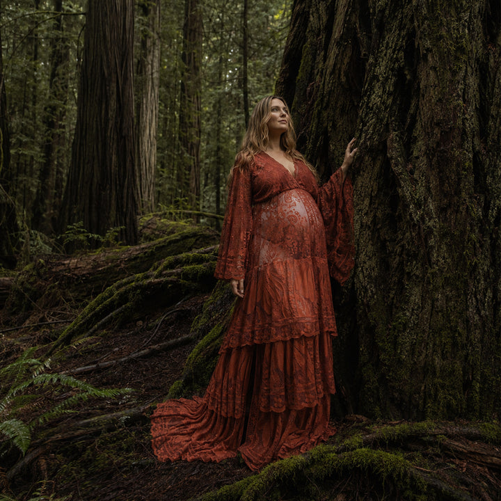 A pregnant woman stands in a dense forest leaning against a large tree trunk covered in moss, wearing a floor length terracotta lace gown.