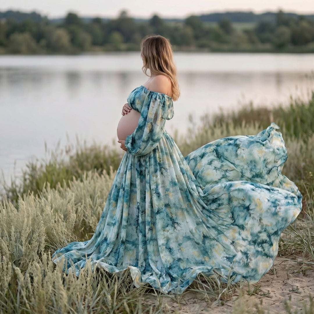 A pregnant woman stands by a lake in a Hestia tie dye print off shoulder dress, holding her belly.
