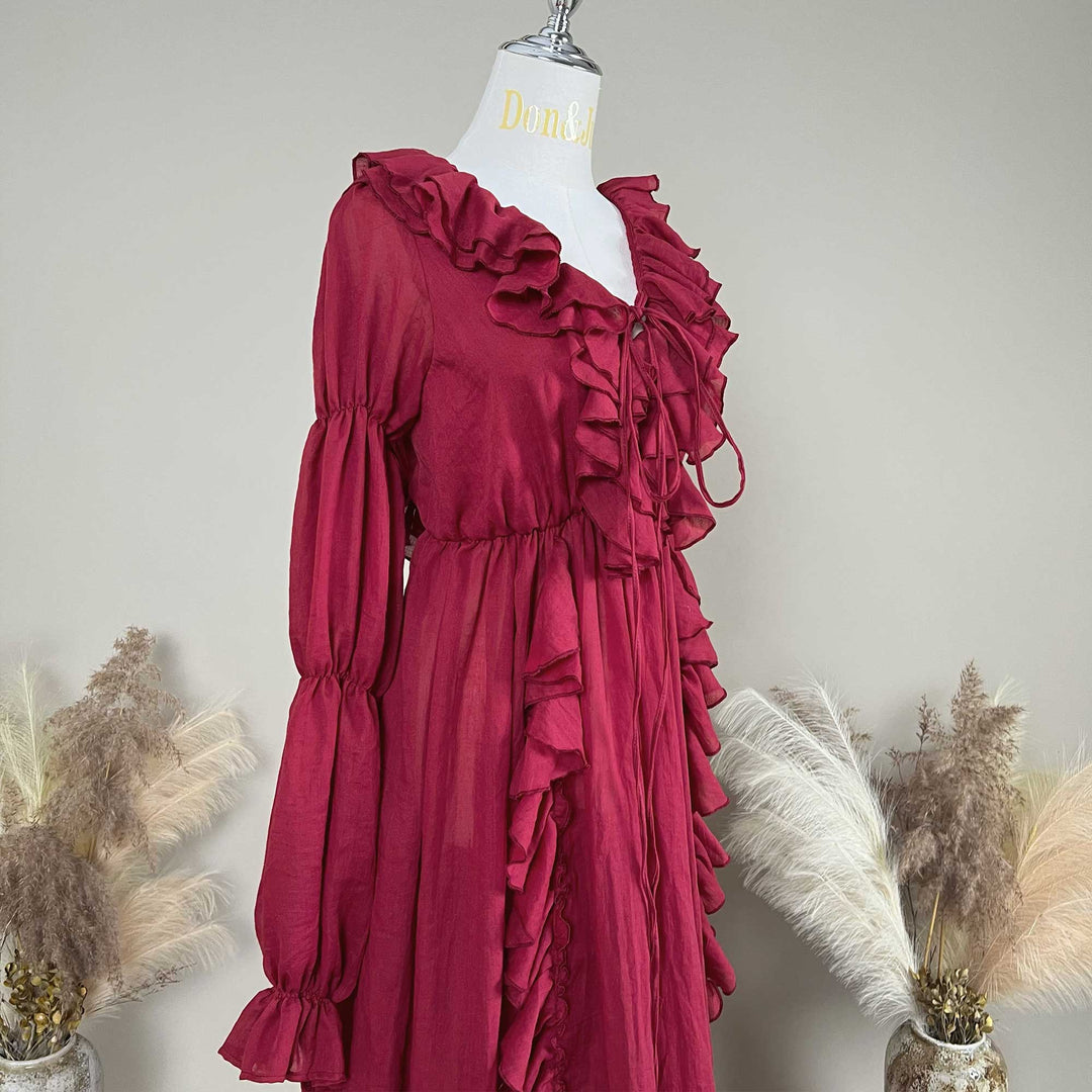 A red Kallectoria dress with dramatic ruffles and long sleeves, displayed on a white mannequin torso. The dress features a tie-front neckline and gathered waist details.