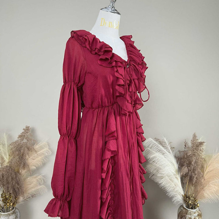 A red Kallectoria dress with dramatic ruffles and long sleeves, displayed on a white mannequin torso. The dress features a tie-front neckline and gathered waist details.