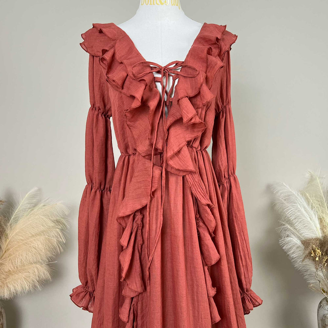 A terracotta-colored linen Kallectoria dress with dramatic ruffles and long sleeves, displayed on a white mannequin torso. The dress features a tie-front neckline and gathered waist details.
