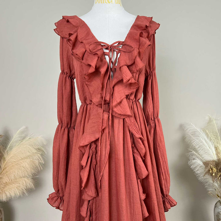 A terracotta-colored linen Kallectoria dress with dramatic ruffles and long sleeves, displayed on a white mannequin torso. The dress features a tie-front neckline and gathered waist details.