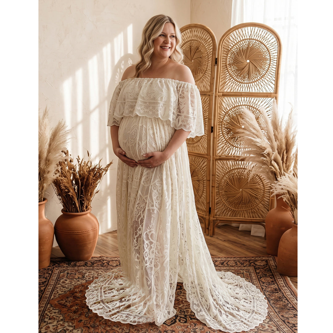 A pregnant woman stands in a beige off-shoulder lace gown holding her belly; the dress features a train, set against a rattan screen, clay vases with dried grass, and a rug.