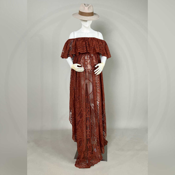 A mannequin displays a terracotta off-shoulder lace gown with ruffled sleeves and a train; the dress features a sheer openwork structure, set against a gray wall with dried flower vases on both sides.