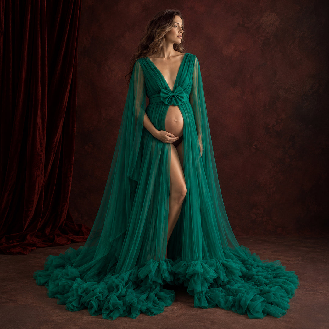A pregnant model wearing a dramatic green sheer tulle maternity gown with a long cape-like drape and ruffled hem, posed against a dark reddish-brown textured backdrop with a red velvet curtain on the left.