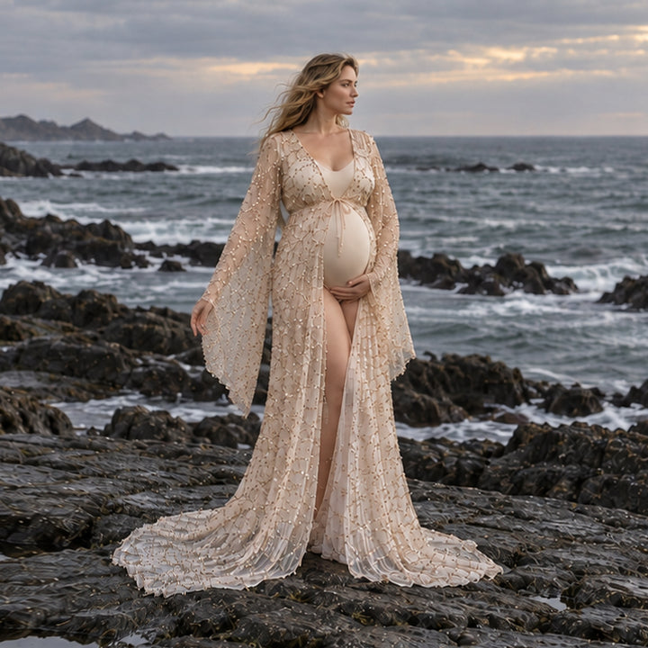 A pregnant woman stands on a rocky ocean shore wearing a long, flowing gown with wide sleeves and a matching headpiece. 