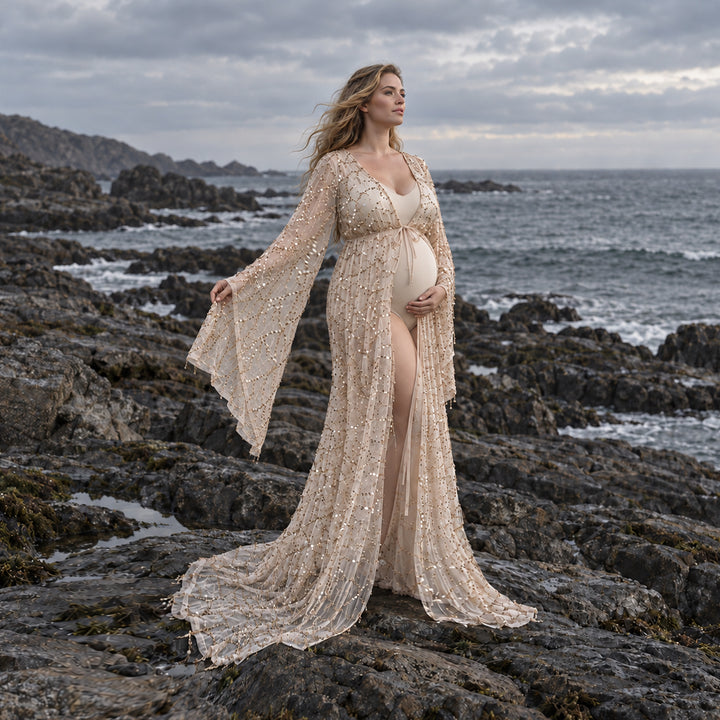 A pregnant woman stands on rocky coastal ground wearing a long, shimmering maternity gown with wide bell sleeves and a sheer overlay, holding her baby bump against an overcast sea and cliff background.