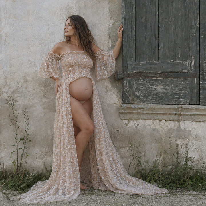 A pregnant woman stands against a rustic textured wall next to an old wooden door, wearing a Lysianassa khaiki floral maternity gown with an open front design.