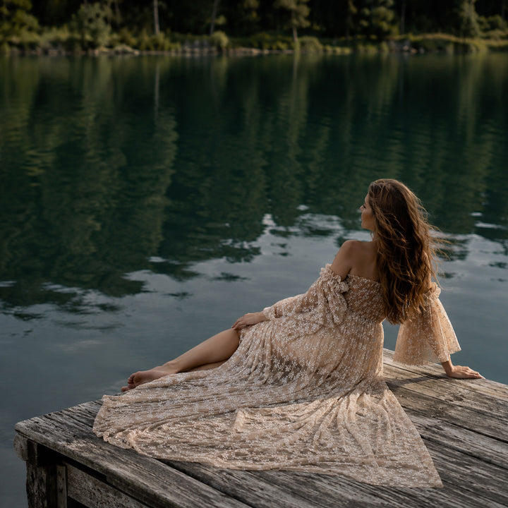 A pregnant woman sits on a weathered wooden dock by a calm lake, wearing a Lysianassa khaiki off-shoulder floral tulle gown. 