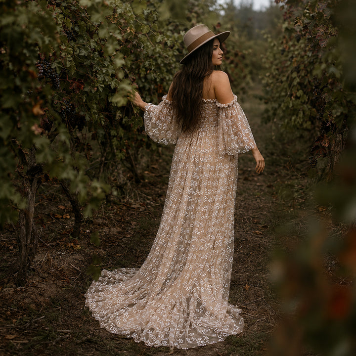 A pregnant woman stands in a vineyard aisle wearing a wide-brimmed hat and a Lysianassa khaiki off-shoulder floral gown. 