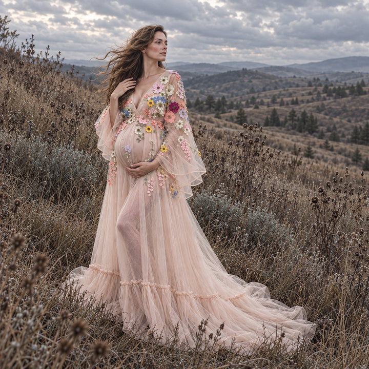 A pregnant woman stands outdoors in a grassy, hilly landscape wearing a flowing tulle gown with colorful floral embroidery and sheer bell sleeves.