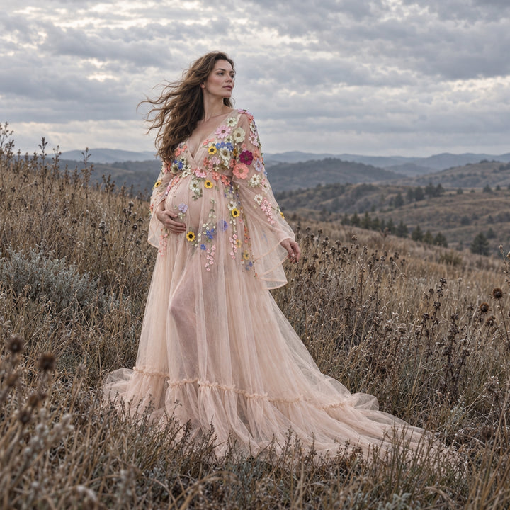 A pregnant woman stands in a grassy field under a cloudy sky, wearing a flowing tulle gown adorned with colorful floral embroidery and long sheer sleeves.