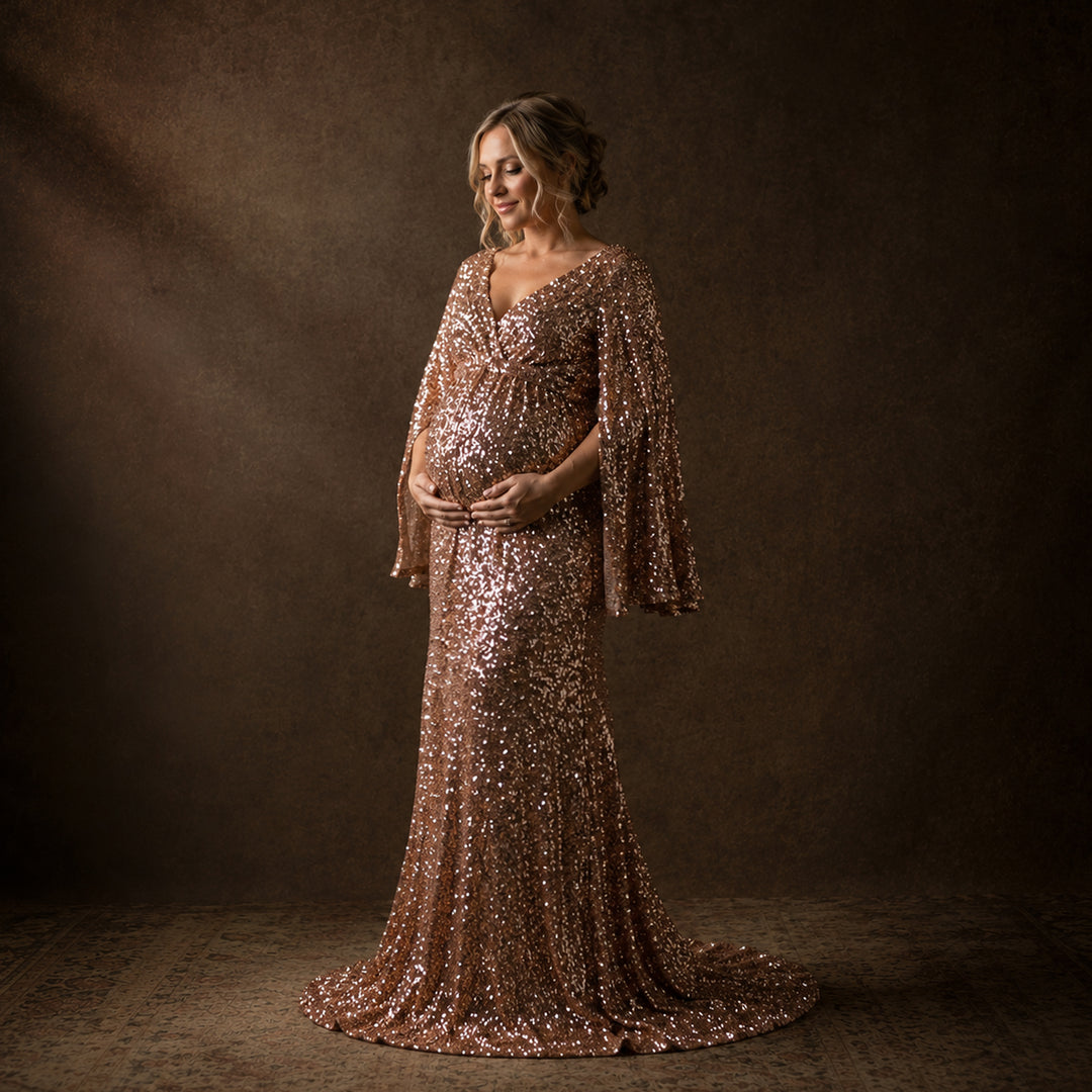 A pregnant woman in a pink gold Mara sequined gown holds her belly with the dress trailing. She stands before a solid-colored backdrop lit from one side.