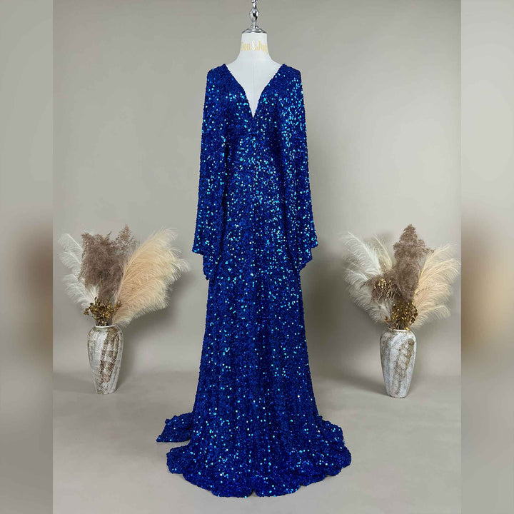 A sapphire blue Mara sequined gown is displayed on a mannequin, featuring a V-neck and flared sleeves. The hem trails on the floor.
