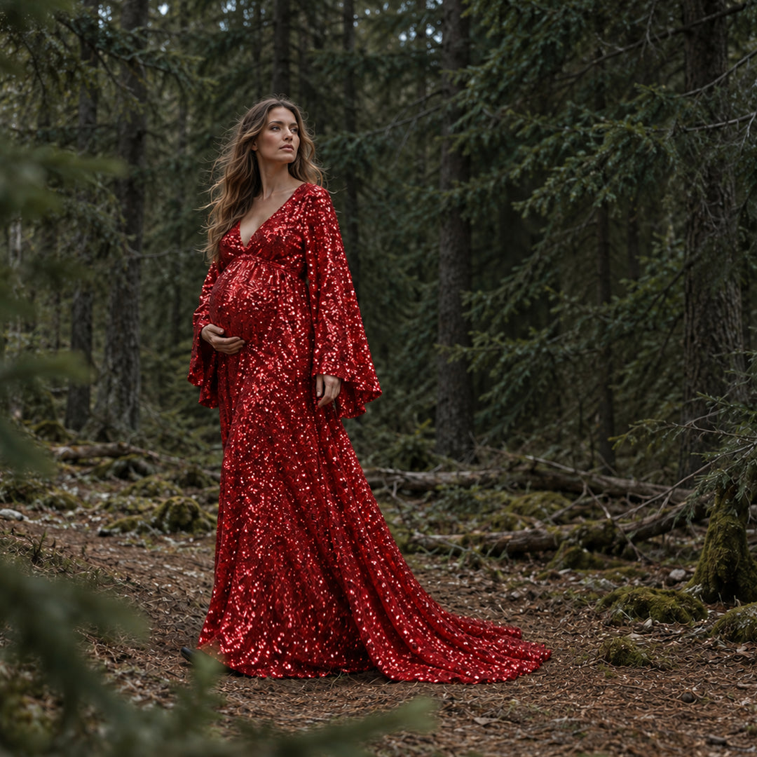 A pregnant woman in a Mara red sequined gown stands in a forest with her left hand on her belly. The hem trails on the mossy ground against dense trees.
