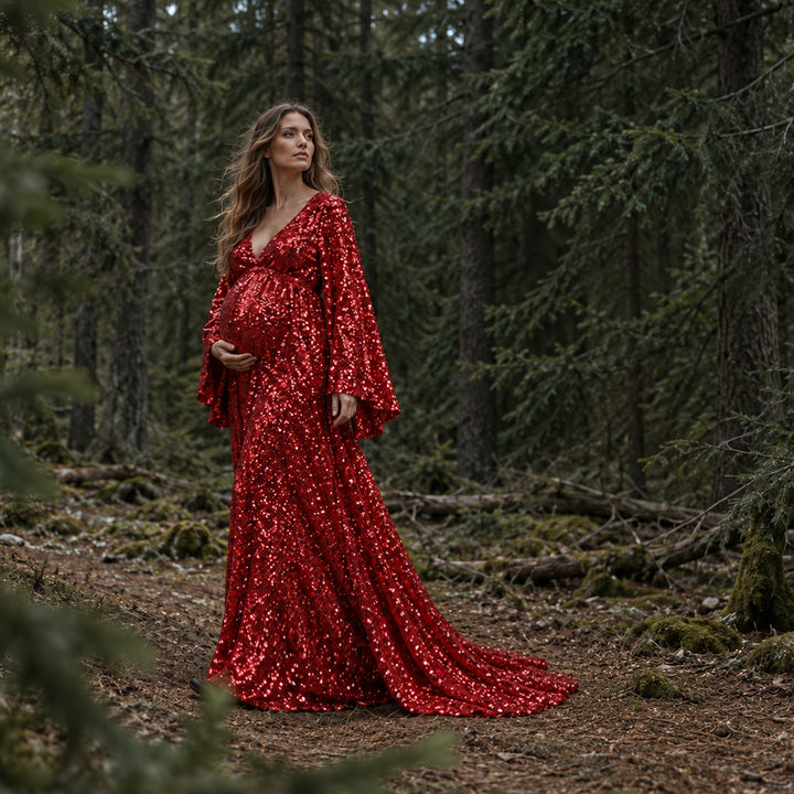 A pregnant woman in a Mara red sequined gown stands in a forest with her left hand on her belly. The hem trails on the mossy ground against dense trees.