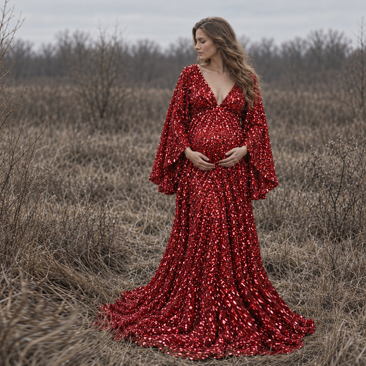 A pregnant woman in a Mara red sequined gown stands in dry grass with both hands on her belly. 