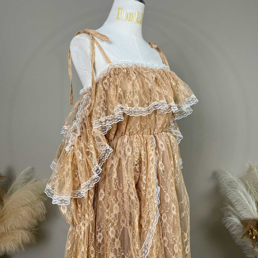 A Melinoe khaki lace maternity dress with dramatic tiered ruffles and off-shoulder sleeves, displayed on a white mannequin torso. The dress features delicate tie-shoulder straps, a gathered elastic waistline, and intricate floral lace fabric trimmed with white scalloped edges.