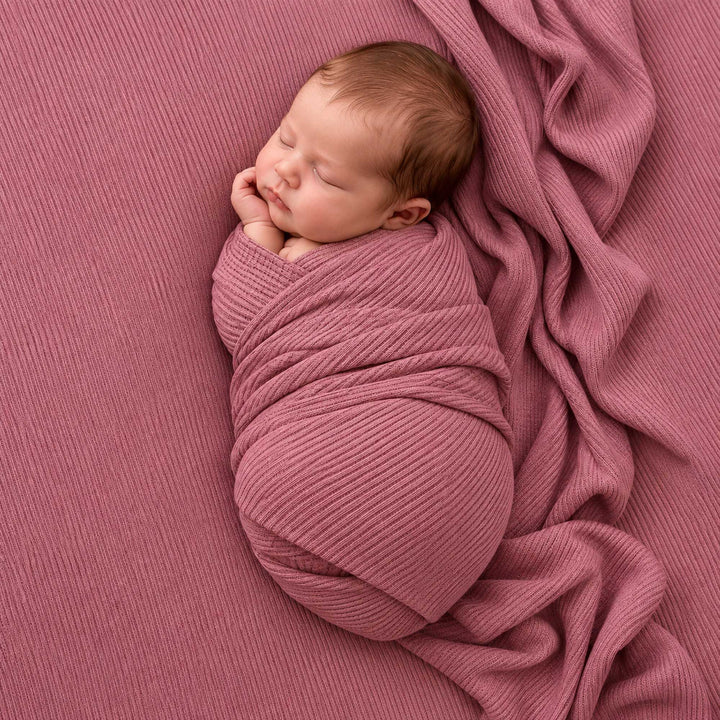 An infant lies on its back on a mauve ribbed knit blanket, body wrapped, hands near the face.