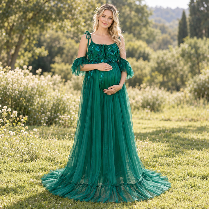 A pregnant woman wears a Oreades green dress with 3D floral embroidery and ruffled sleeves stands on the grass holding her belly.