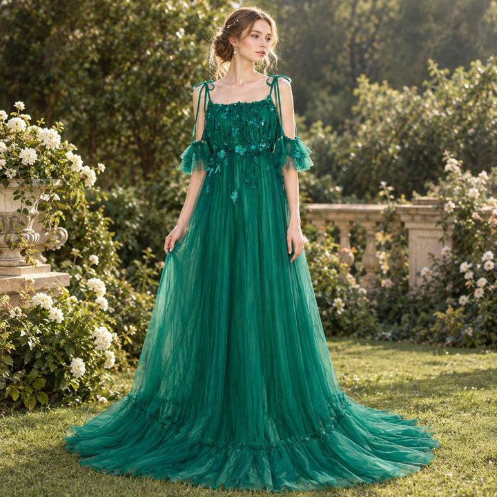 A woman stands in a garden wearing a Oreades green tulle gown with tie straps and ruffled sleeves. Green bushes and white flowers are visible in the background behind her.
