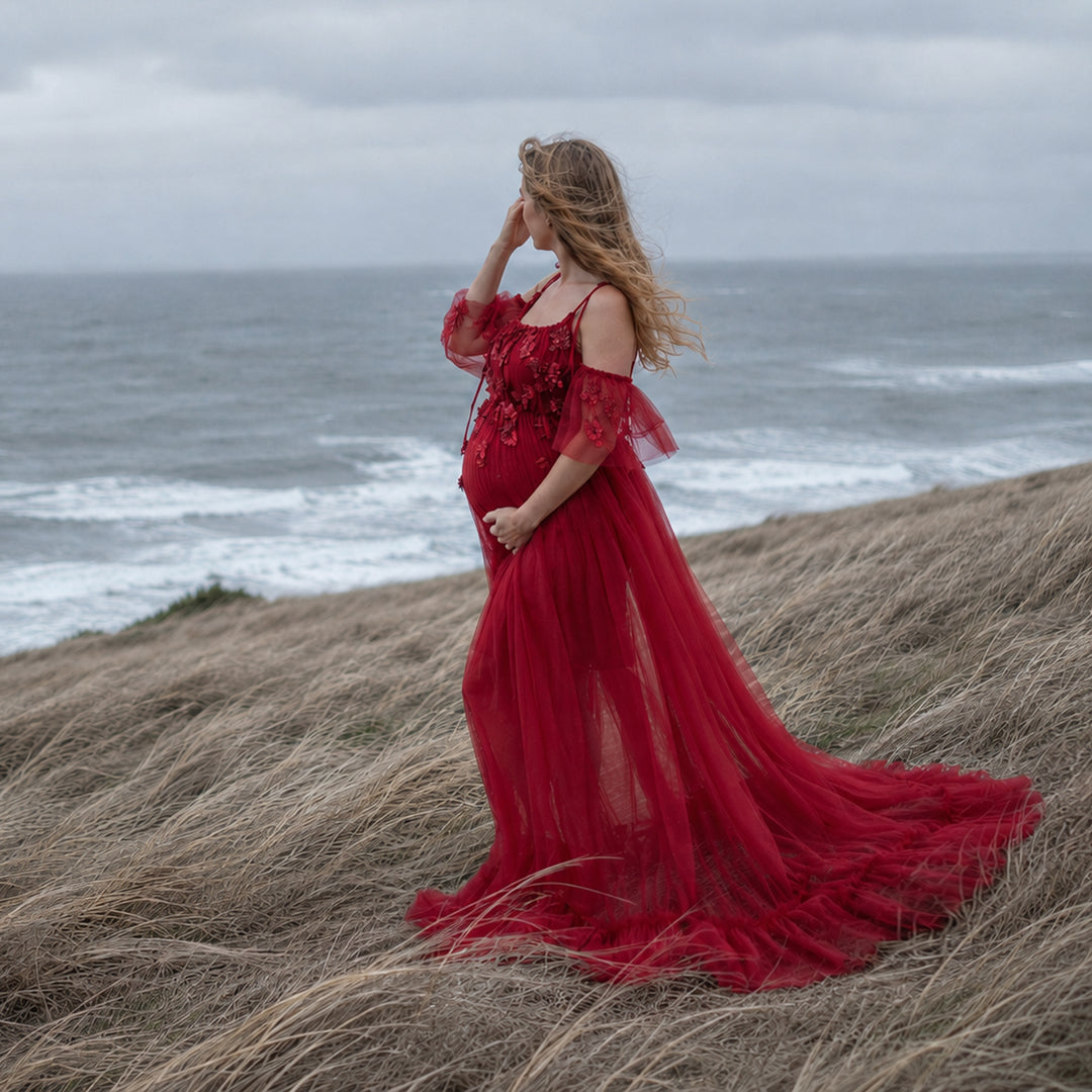 A pregnant woman in a Oreades red spaghetti-strap gown with a trailing skirt stands on a grassy slope; she touches her hair with her right hand, cradles her belly with her left, with the sea and waves in the background.