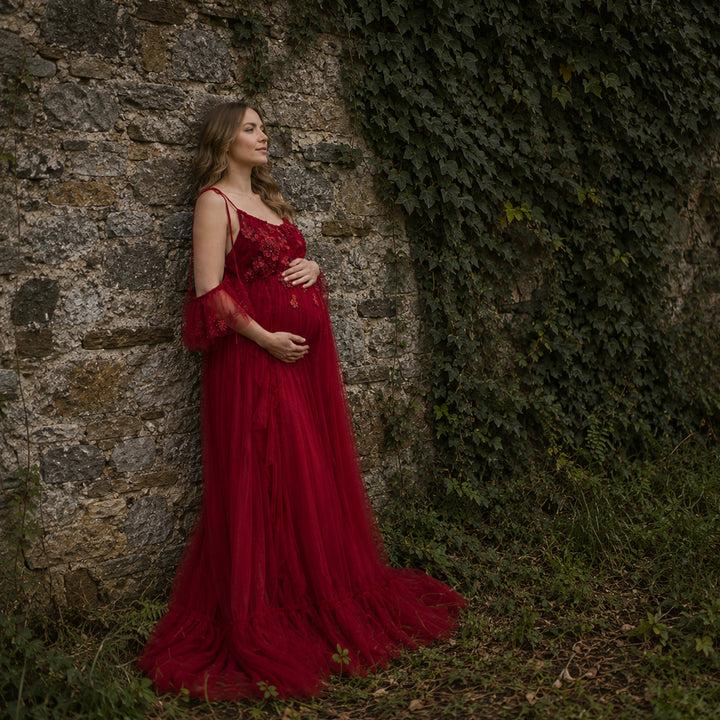  pregnant woman in a red spaghetti-strap gown with floral embroidery, ruffled sleeves and a trailing skirt stands before a wooden cabin; she cradles her belly with both hands, looking down.