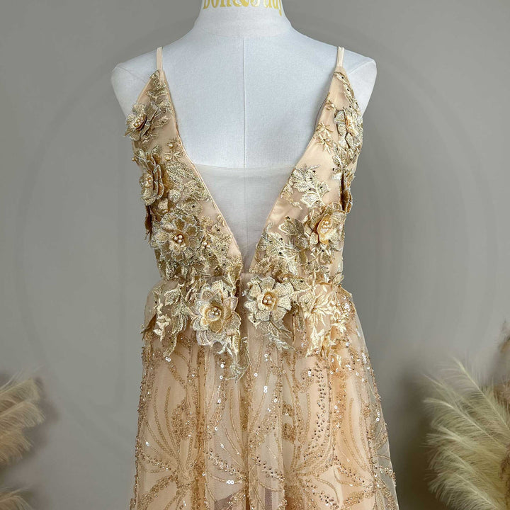A mannequin displays the upper body of a Pandia gold spaghetti strap gown with a deep V-neck and 3D floral decorations.