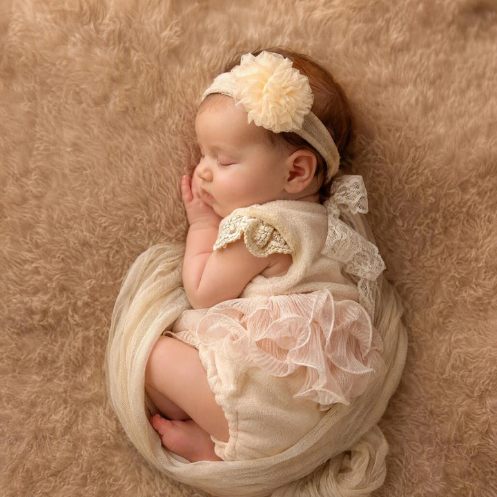 A newborn who wears a skin color romper featuring crocheted ruffled short sleeves lies on its side on a blanket.