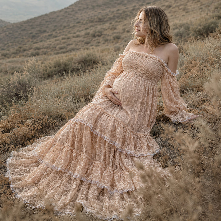 A pregnant woman in a khaki Selene off-shoulder lace dress sits on a hill covered in dry plants, holding her baby bump.