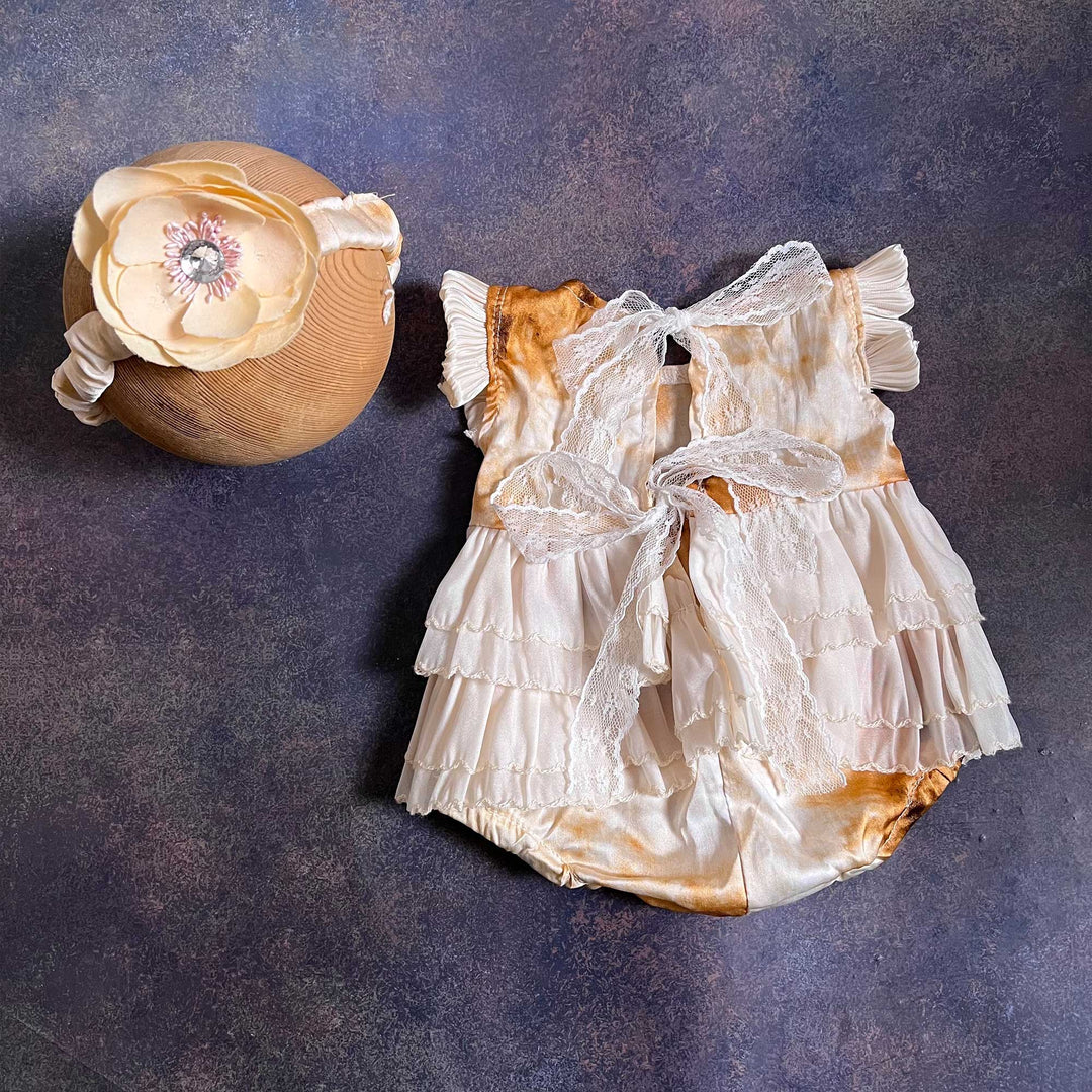 A romper shown from the back, with a white lace ribbon bow and tiered skirt, a flower headband placed on the left.