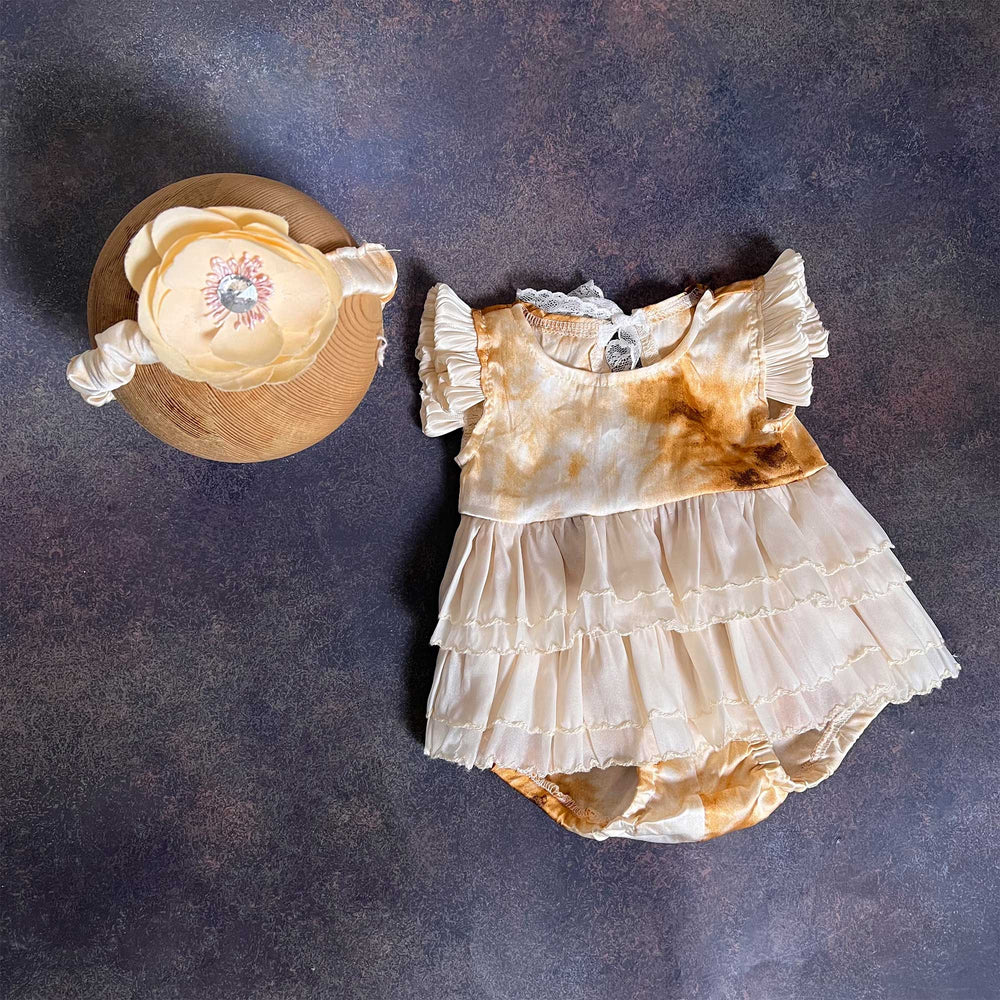 A romper laid flat from the front, showing the yellow tie-dye bodice, ruffled sleeves and three-tier skirt, with the same flower headband on the left.