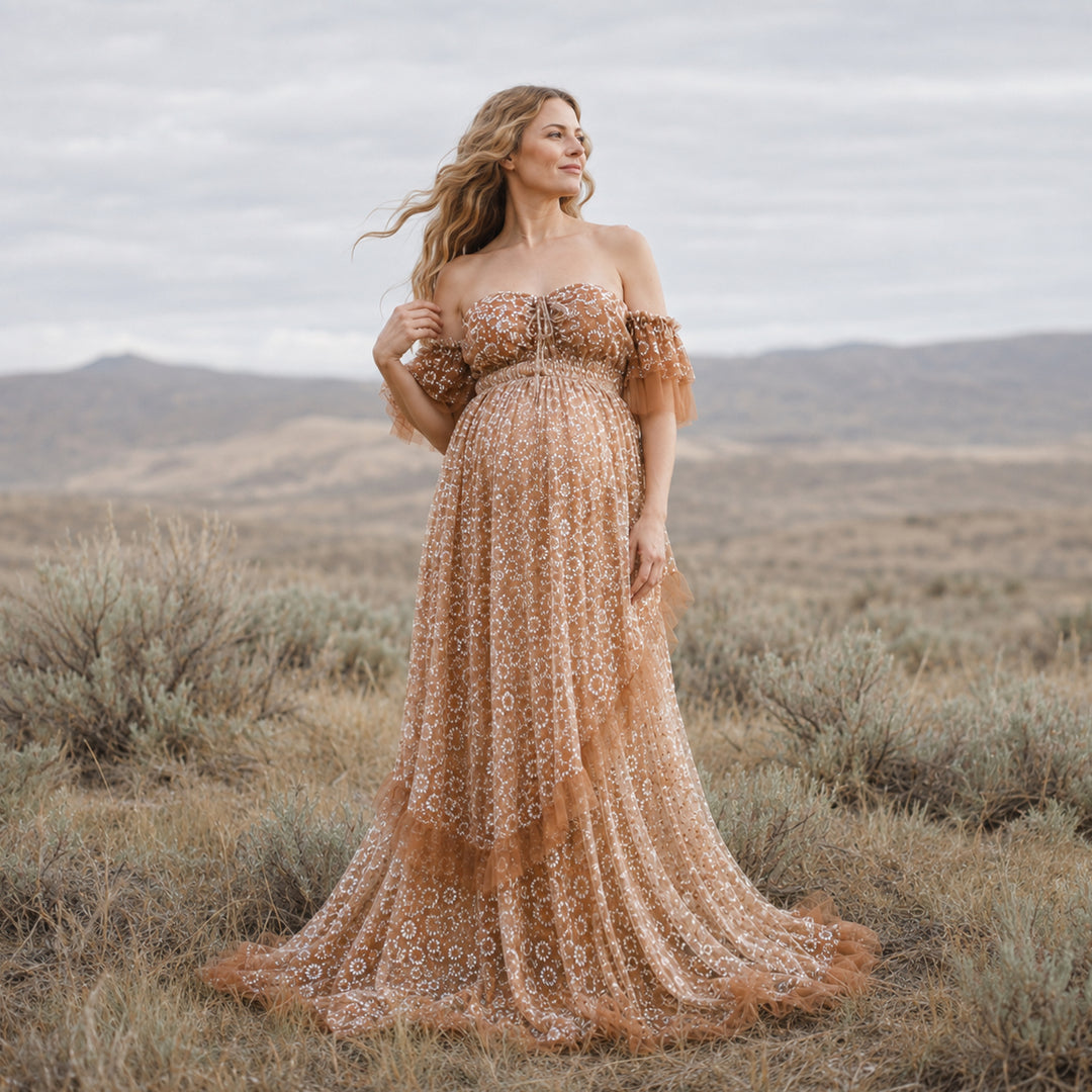 A woman is wearing Tyche khaki sheer off-shoulder gown stands outdoors, the dress featuring white embroidery; she touches her shoulder with her right hand.
