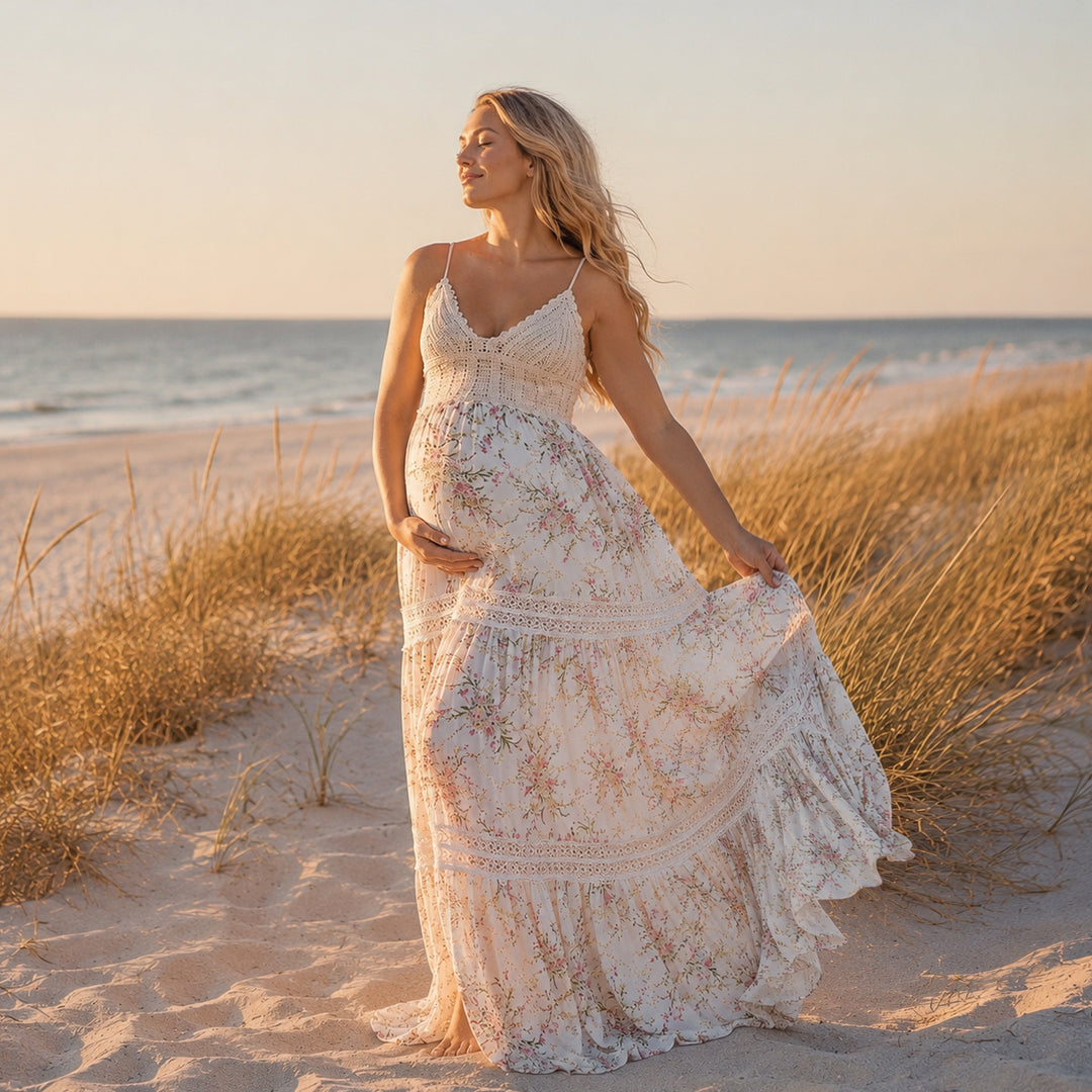 A pregnant woman stands on a sandy beach wearing an apricot bottom floral maternity maxi dress with spaghetti straps, a crochet bodice, and a tiered skirt with lace inserts.