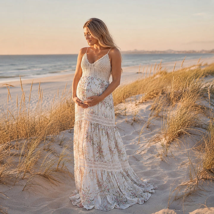 A pregnant woman stands on a sandy beach wearing an apricot bottom floral  maternity maxi dress with spaghetti straps, a crochet bodice, and a tiered skirt with lace inserts.