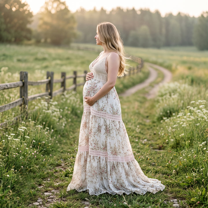 A pregnant woman stands in profile on a grassy path next to a wooden fence, wearing an apricot bottom floral print maternity maxi dress with a crochet bodice, spaghetti straps, and lace-trimmed tiered panels on the skirt.
