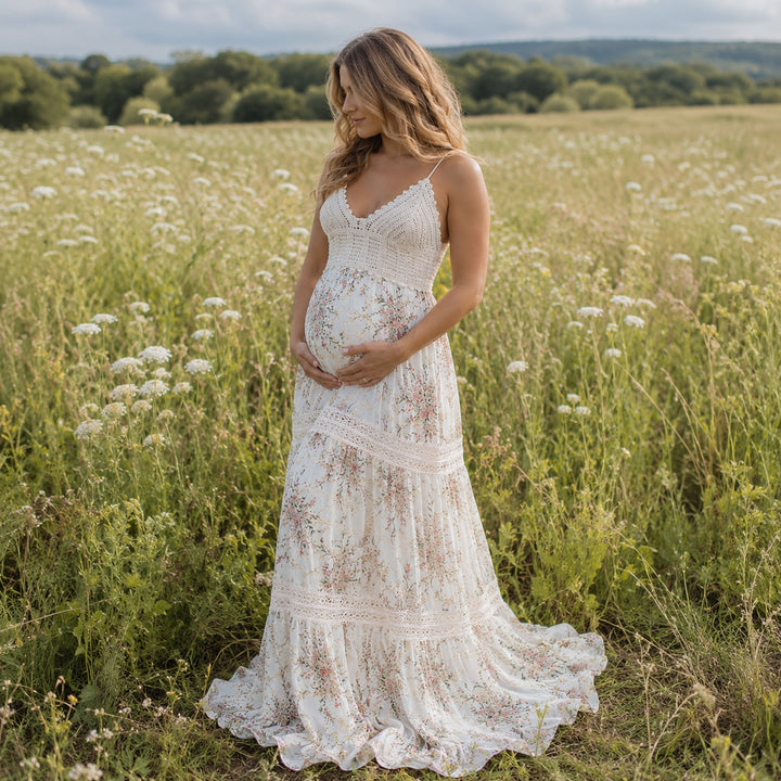 A pregnant woman stands in a field of tall grass and white wildflowers wearing an apricot bottom floral print maternity maxi dress with spaghetti straps, a crochet bodice, and lace-trimmed tiered panels on the skirt that pools on the ground.